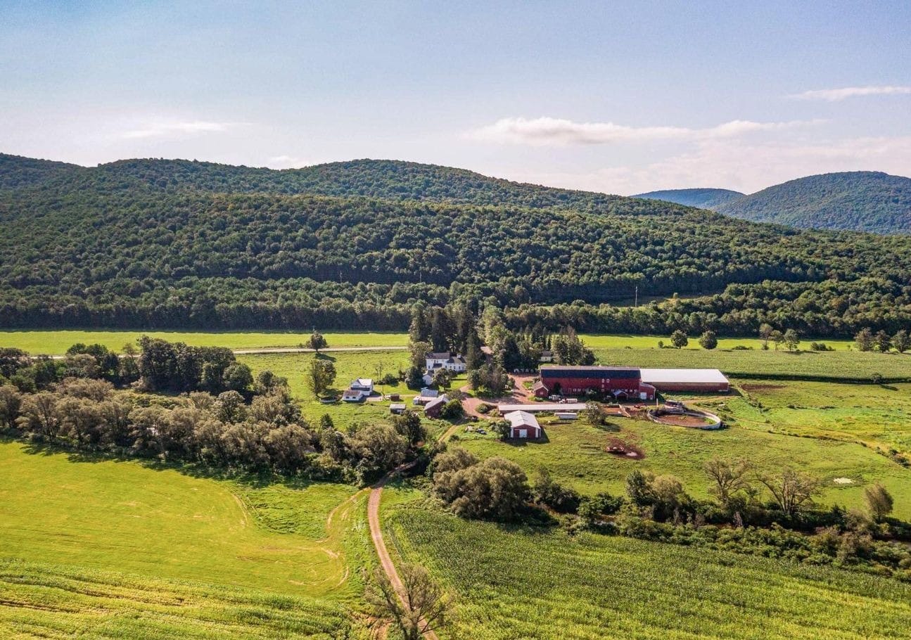 Ashridge farm campus — aerial view of the Greek Revival farmhouse, red barn complex, fields, and Catskill hills, South Kortright, New York