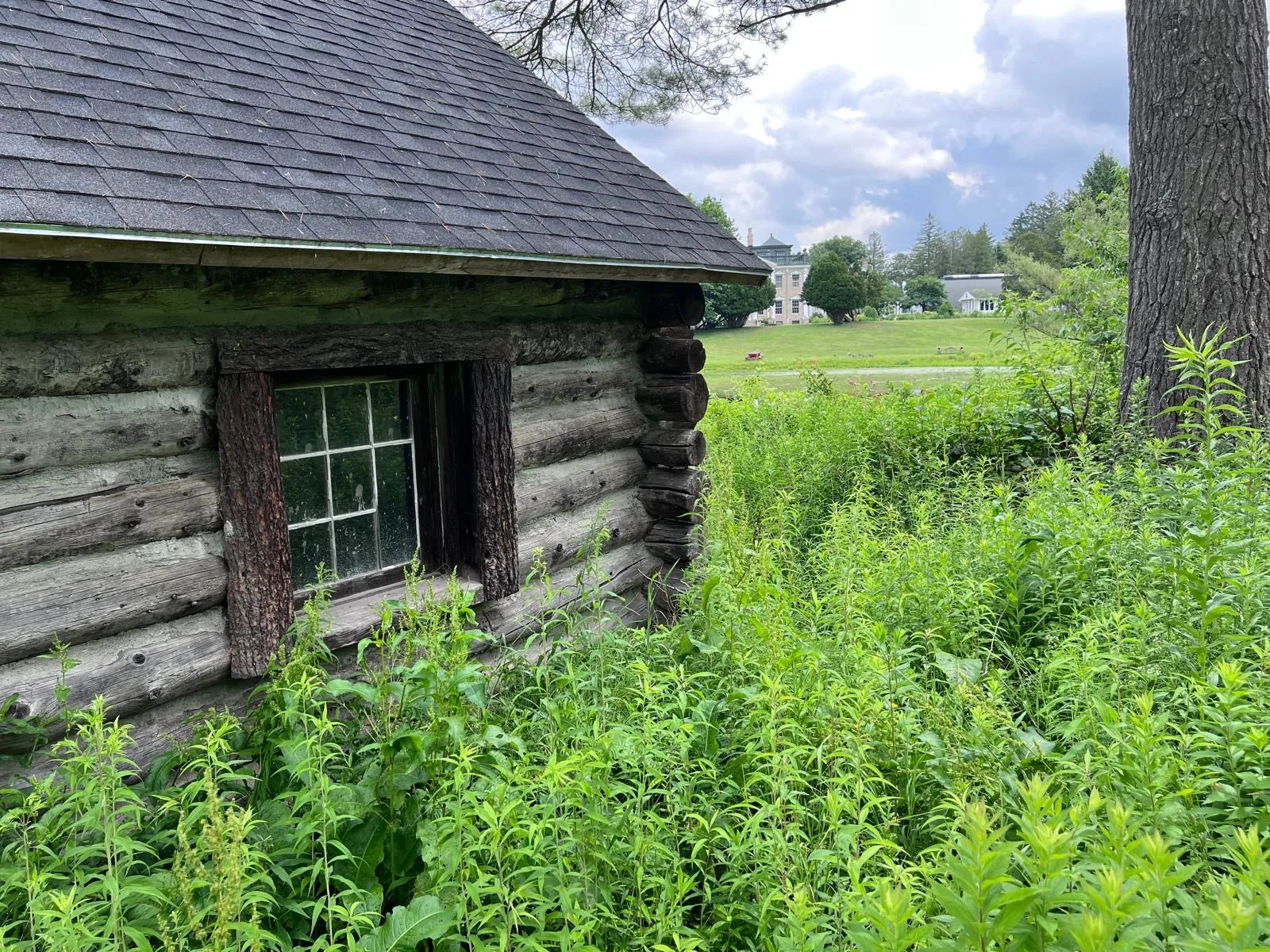 Exterior corner of the log boathouse with its shingled roof, the estate mansion visible across the lawn beyond.