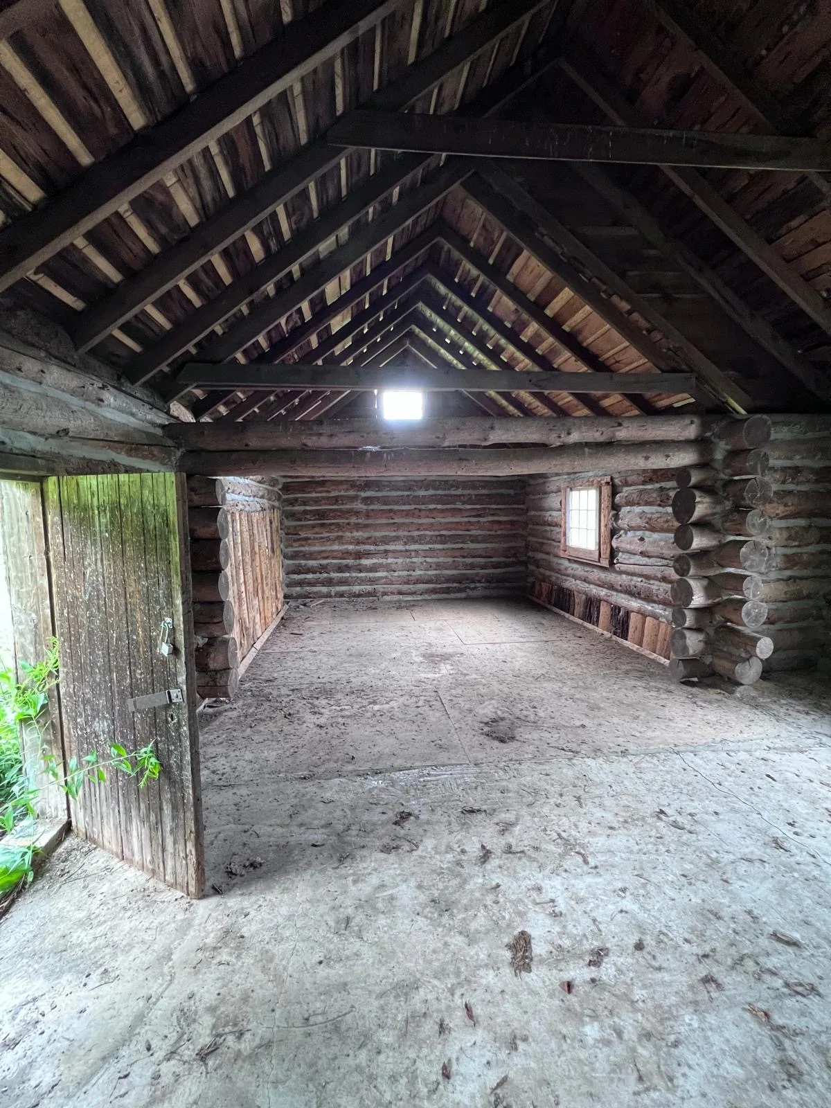 Interior of the original log boathouse — exposed round-log walls and a timber cathedral roof, the plank door open to the lakeshore.