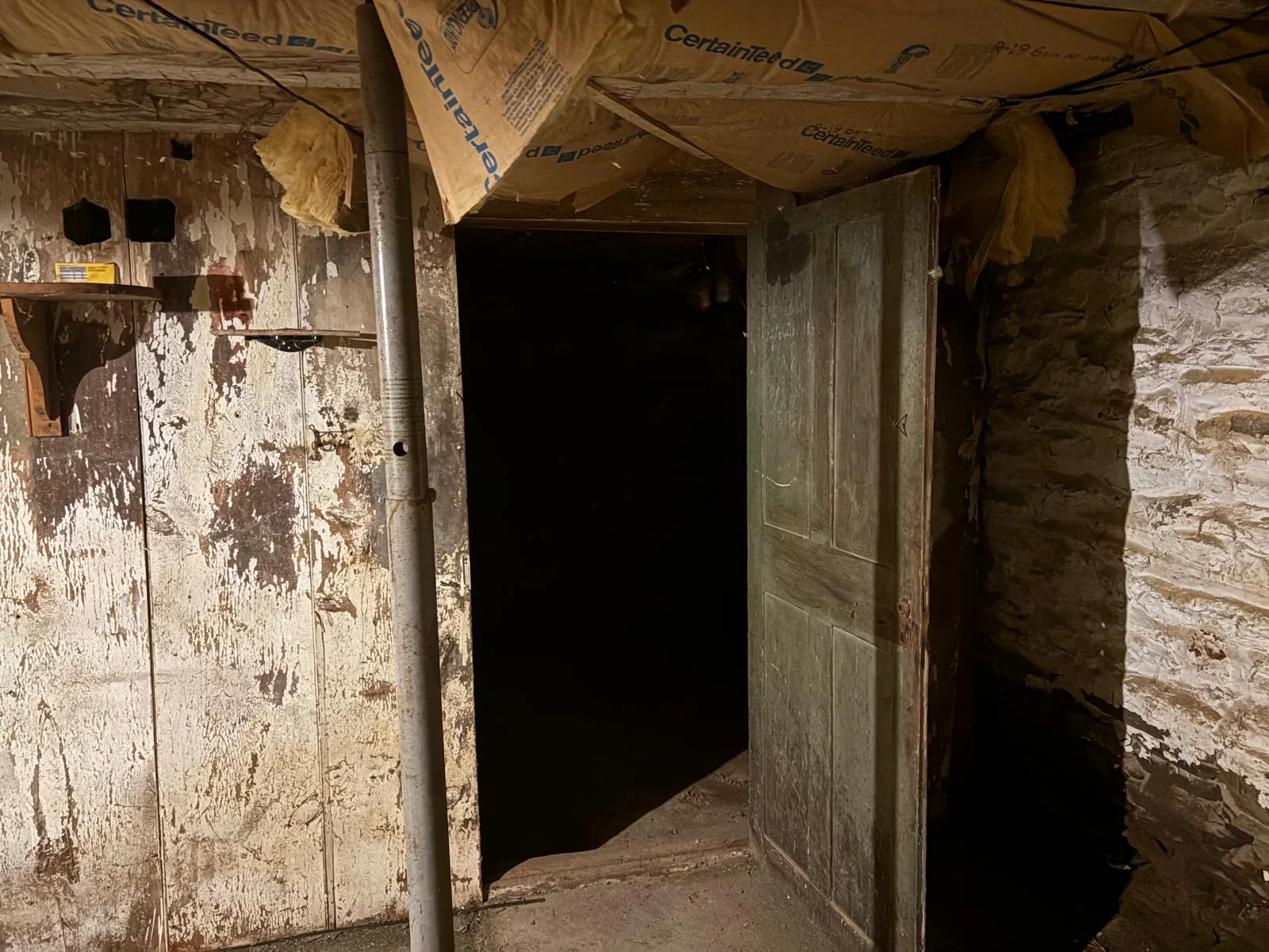 Original paneled door opening into a dark stone cellar room — peeling paint and whitewashed stone.