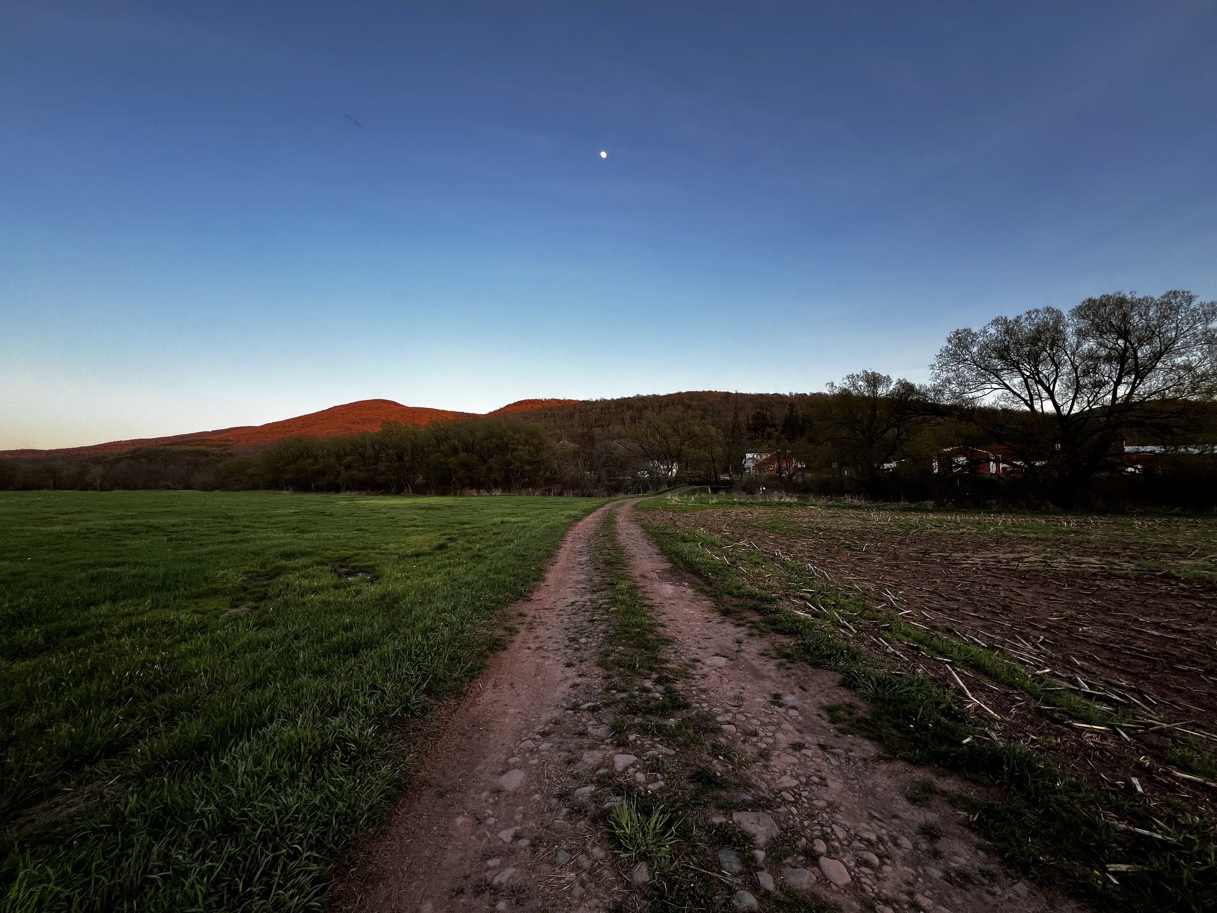 Ashridge farm acreage at dusk — field road, red barns, and last light on the Catskill ridge, South Kortright, New York