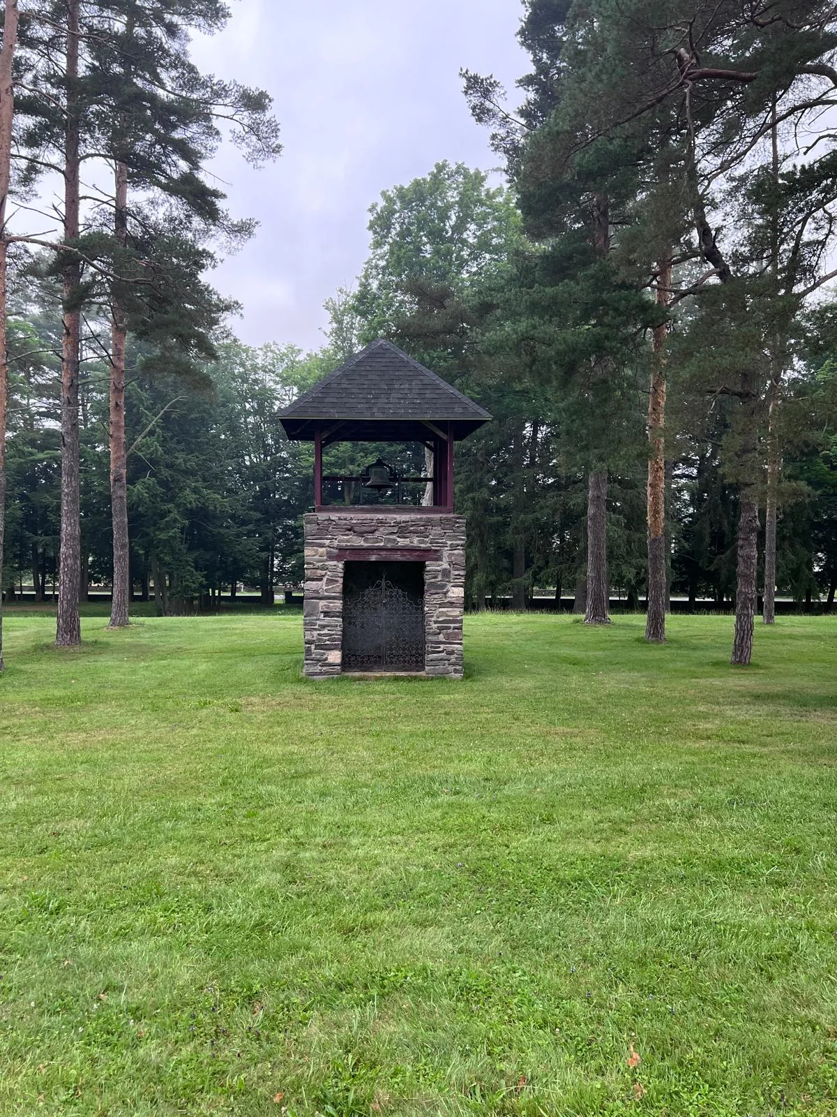 The estate's original stone-and-timber bell tower — a bell in an open timber belfry over an iron-gated stone base, standing on lawn in a grove of tall pines.