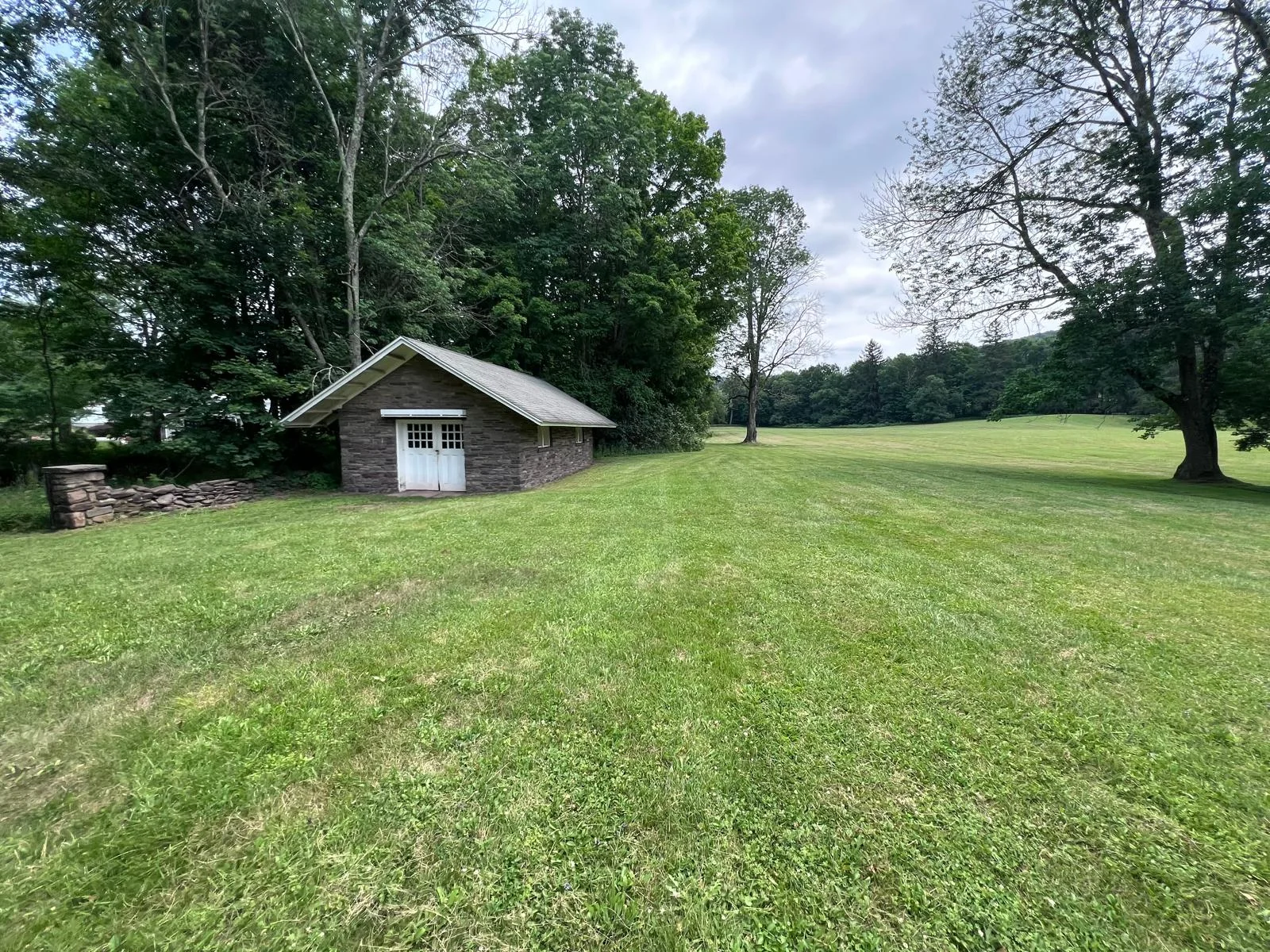 Wide view of the stone house across open lawn — white carriage doors and a dry-laid stone wall at the treeline.
